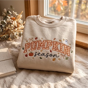 Puede incluir: Una sudadera de cuello redondo en color beige con texto bordado que dice PUMPKIN season en letras mayúsculas de cuadros naranjas y blancos y en cursiva negra. El diseño incluye pequeñas hojas de otoño bordadas, una calabaza y un fantasma.