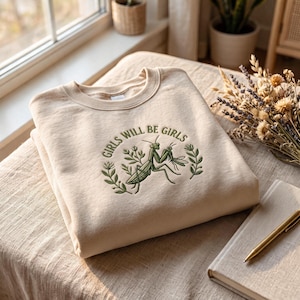 Può includere: Una felpa girocollo beige con un ricamo verde raffigurante una mantide religiosa al centro del petto. Il design include dettagli botanici a foglia e il testo "GIRLS WILL BE GIRLS" ricamato ad arco in filo verde scuro sopra l'insetto.