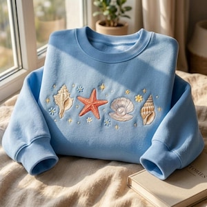 May include: A light blue sweatshirt with embroidered seashells, a starfish, and a pearl. The design includes small flowers and stars. The sweatshirt has a crew neck and long sleeves.