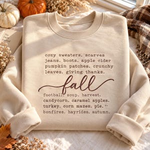 Pode incluir: Uma camisola bege de gola redonda com texto bordado em castanho escuro que lista palavras com o tema de outono, incluindo cozy sweaters, scarves, jeans, boots, apple cider, pumpkin patches, crunchy leaves, giving thanks, fall, football, soup, harvest, candycorn, caramel apples, turkey, corn mazes, pie, bonfires, hayrides e autumn.