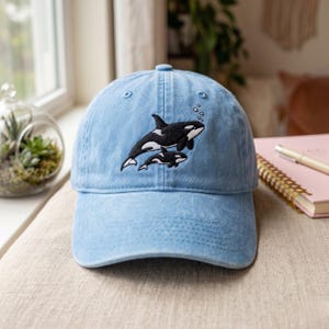Könnte beinhalten: Eine hellblaue Baseballkappe mit einer Stickerei, die einen großen Orca über einem kleineren Orca-Kalb zeigt. Das Motiv ist in schwarzem und weißem Garn gehalten, ergänzt durch kleine weiße Kreise, die aufsteigende Luftblasen darstellen.