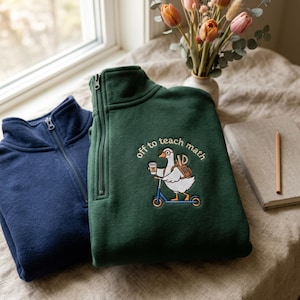 Pode incluir: Uma camisola verde-escura com fecho de um quarto, com um bordado de um ganso num patinete azul, segurando um copo de café com o símbolo pi, usando óculos e uma mochila, com o texto off to teach math, ao lado de uma camisola azul-marinho.