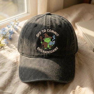 May include: A dark gray baseball cap with the phrase "OFF TO COMMIT SHENANIGANS" embroidered on the front. The design features a cartoon frog wearing a wizard hat and holding a book. The cap is made of a soft, worn fabric.