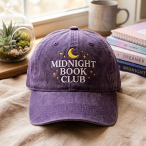 Könnte beinhalten: Eine violette Baseballkappe mit weißem Sticktext, der MIDNIGHT BOOK CLUB lautet, zentriert unter einem gelben Halbmond und kleinen Sternsymbolen. Die Kappe hat eine strukturierte Krone, einen gebogenen Schirm und sichtbare Nähte.