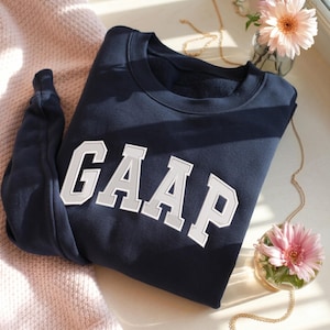 Può includere: Una felpa blu navy a girocollo con la scritta GAAP in grandi lettere bianche in stile blocco con un contorno grigio chiaro. Il capo presenta maniche lunghe, un colletto a coste e una fodera morbida, con un design pulito ed essenziale.