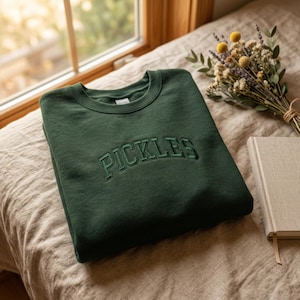 Könnte beinhalten: Ein dunkelwaldgrünes Sweatshirt mit Rundhalsausschnitt, auf dessen Brust das Wort PICKLES in passendem dunkelgrünem Garn in einer geschwungenen, im College-Stil gehaltenen Schriftart gestickt ist. Das Kleidungsstück hat lange Ärmel.