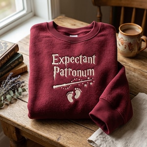 May include: A burgundy crewneck sweatshirt with the embroidered text "Expectant Patronum" in a stylized font. Below the text is a wand, stars, and baby footprints. The sweatshirt is folded on a wooden table.