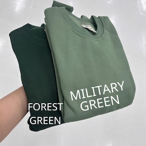 May include: Two crewneck sweatshirts featuring a solid color design. One sweatshirt is a dark forest green, and the other is a lighter military green. The text FOREST GREEN is printed near the dark sweatshirt, and MILITARY GREEN is near the light one.