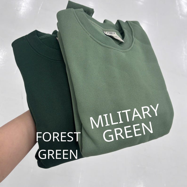 May include: Two crewneck sweatshirts featuring a solid color design. One sweatshirt is a dark forest green, and the other is a lighter military green. The text FOREST GREEN is printed near the dark sweatshirt, and MILITARY GREEN is near the light one.