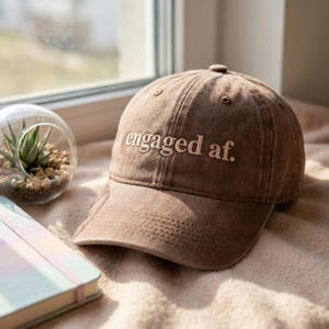 May include: A brown baseball cap with the embroidered text "engaged af." The hat is made of a soft, textured material. A small succulent terrarium and a notebook are visible in the background.