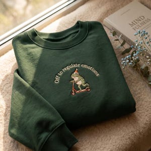 Puede incluir: Una sudadera de cuello redondo verde oscuro con un bordado de una rana con gorro de Papá Noel rojo y blanco montada en un patinete rojo. El texto "Off to regulate emotions" aparece en un semicírculo sobre la ilustración de la rana.