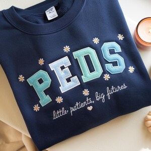 May include: A navy blue crewneck sweatshirt featuring the embroidered text PEDS in large, light blue and teal varsity-style letters. The design includes small white daisy flowers and the cursive text little patients, big futures.