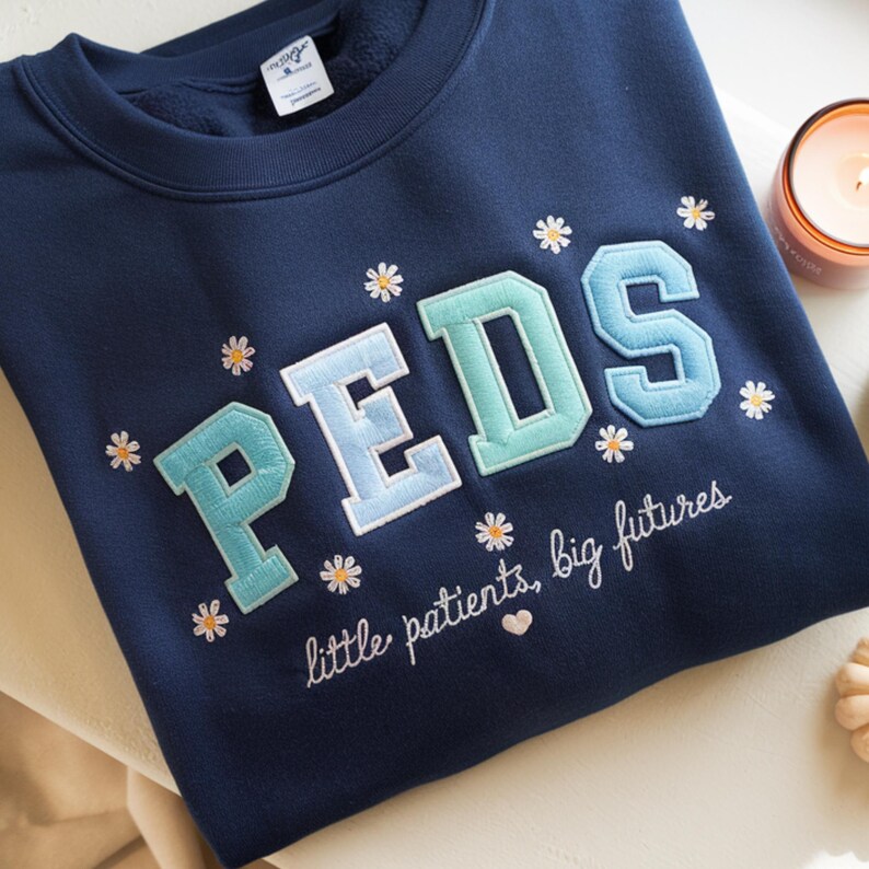May include: A navy blue crewneck sweatshirt featuring the embroidered text PEDS in large, light blue and teal varsity-style letters. The design includes small white daisy flowers and the cursive text little patients, big futures.