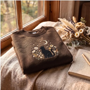Könnte beinhalten: Ein dunkelbraunes Sweatshirt mit Rundhalsausschnitt und einer Stickerei, die eine schwarze Katze inmitten von weißen und gelben Blumen zeigt, darüber eine Mondsichel und kleine Sterne. Die Stickerei befindet sich mittig auf der Brust.