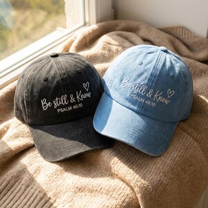 May include: Two baseball caps, one in charcoal grey and one in light blue, both featuring a distressed fabric finish. Each cap is embroidered with the white text "Be still & Know" followed by "PSALM 46:10" and a small heart outline symbol.