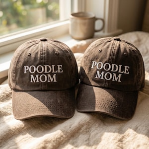 May include: Two dark brown baseball caps with a distressed finish, each featuring the white embroidered text POODLE MOM on the front panel. The caps have a structured crown, a curved brim, and stitched eyelets for ventilation on the upper panels.
