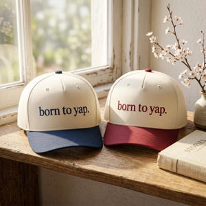 Peut inclure: Deux casquettes de baseball avec des calottes couleur crème et des panneaux structurés. L'une présente une visière bleu foncé et le texte "born to yap." brodé en bleu foncé. L'autre a une visière bordeaux et le texte "born to yap." en bordeaux.