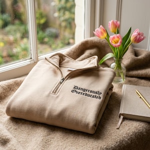 May include: A beige quarter-zip sweatshirt featuring a high collar with a metal zipper. The chest area displays the text "Dangerously Overeducated" embroidered in a black gothic-style font. The garment is made of a solid-colored fabric.