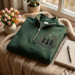 Puede incluir: Una sudadera verde oscuro con cremallera corta que presenta un diseño bordado de tres figuras con sombreros puntiagudos bailando bajo una luna creciente y árboles, con el texto "GIRLS WILL BE GIRLS" cosido en línea curva bajo las figuras.
