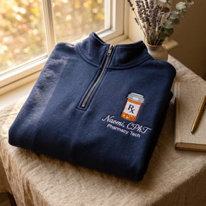 May include: A navy blue quarter-zip sweatshirt featuring an embroidered graphic of an orange prescription pill bottle with an Rx symbol. Below the bottle, white text reads "Naomi, CPhT" in a script font and "Pharmacy Tech" in a sans-serif font.