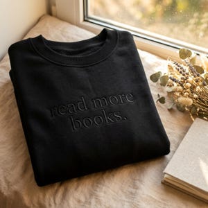Może przedstawiać: Czarna bluza z haftowanym napisem "read more books". Ubranie jest złożone na beżowej powierzchni, blisko okna. Widoczna jest również książka i suszone kwiaty.