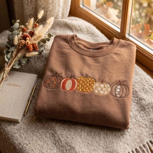 Puede incluir: Una sudadera marrón de cuello redondo con una fila horizontal bordada de cinco calabazas en el pecho. Cada calabaza muestra un patrón único, incluyendo motivos florales, rayas verticales, lunares y diseños de margaritas en tonos naranja, blanco y marrón.