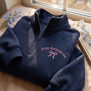 May include: A navy blue quarter-zip sweatshirt featuring the embroidered phrase "in my doctorate era" and a pink bow. The sweatshirt has a high collar and a silver zipper. The garment is made of a soft material.