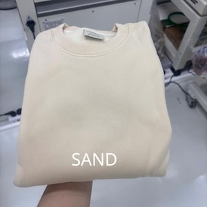 May include: A solid light beige crewneck sweatshirt with a ribbed collar and cuffs. The word SAND is printed in white capital letters on the lower center of the garment. A small rectangular fabric label is visible inside the neckline.