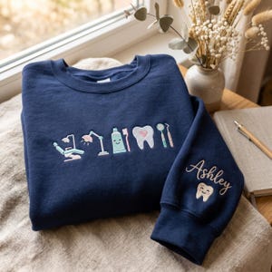Op de afbeelding: Marineblauwe sweatshirt met geborduurde tandheelkundige ontwerpen: een tandartsstoel, lamp, tandpasta, tandenborstel, tand met een hart en tandheelkundige instrumenten. De rechter mouw toont de naam "Ashley" en een lachende tand.