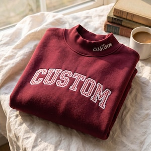 May include: A deep burgundy sweatshirt featuring a mock neck collar with the word custom embroidered in white. The front displays the word CUSTOM in large, arched block letters with a white and red gingham checkered pattern fill.