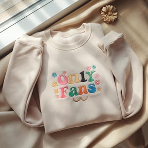 Pode incluir: Camisola bege com a frase bordada "Only Fans" em letras coloridas e detalhes florais. A parte superior de manga comprida e gola redonda é feita de um material macio.
