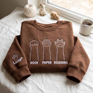 Op de afbeelding: Een bruine sweater met ronde hals, voorzien van witte geborduurde kattenpootjes en de tekst "ROCK PAPER SCISSORS" op de voorzijde. De linkermouw bevat een geborduurde pootafdruk en de naam "Luna" met een klein hartje.