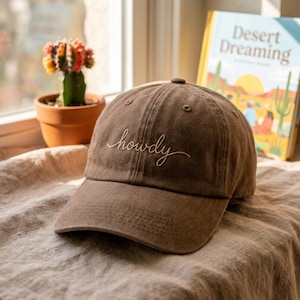 Op de afbeelding: Bruine baseballcap met het woord "howdy" geborduurd in wit script. De cap ligt op een neutraal gekleurd oppervlak, met een cactus in een terracotta pot en een boek met de titel "Desert Dreaming" op de achtergrond.