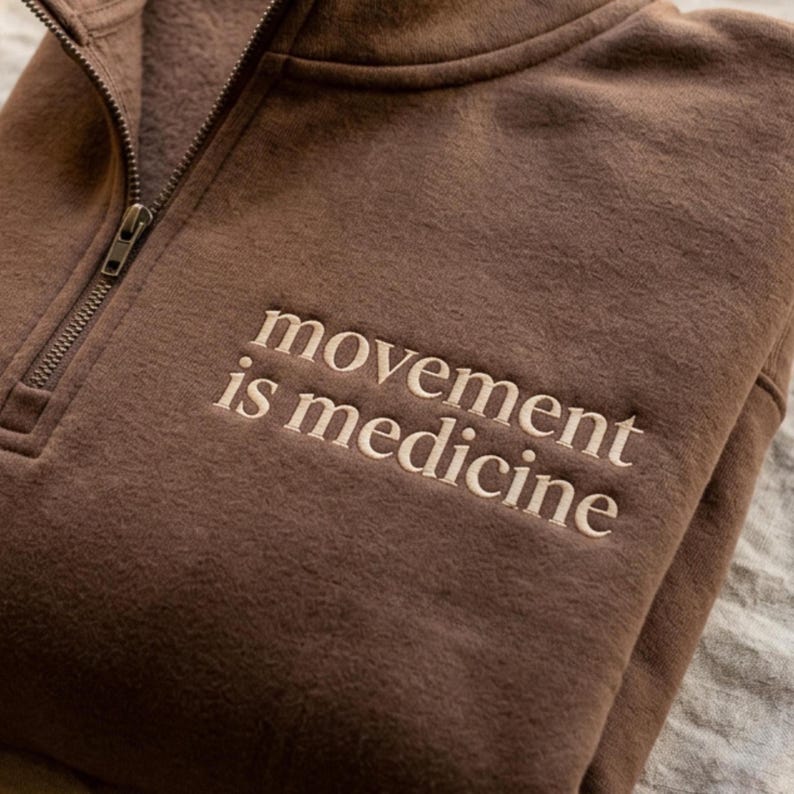 May include: A brown fleece pullover with a quarter-zip neckline. The phrase "movement is medicine" is embroidered in off-white thread on the front. The fabric appears soft and comfortable.