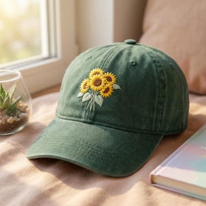Op de afbeelding: Een groene baseballcap met een geborduurd boeket zonnebloemen. De zonnebloemen hebben gele bloemblaadjes en bruine centra. De cap is gemaakt van zachte stof en heeft een gebogen klep.