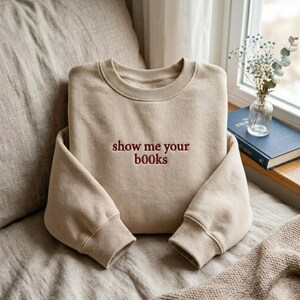 May include: A beige sweatshirt with the embroidered phrase "show me your books" in burgundy lettering. The sweatshirt has a crew neck and ribbed cuffs and hem. The image is taken indoors, with a window and a book in the background.
