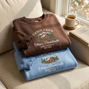 May include: Two crewneck sweatshirts in brown and light blue, each featuring an embroidered graphic of a cabin, mountains, and pine trees with the text CABIN CREW, Olivia's Bachelorette, LAKE TAHOE, CA, EST. 2025 stitched in white thread.
