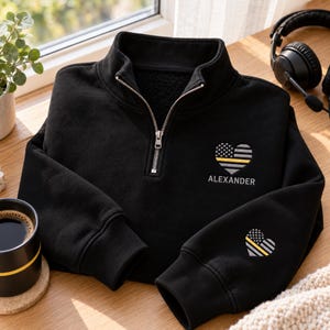 May include: A black quarter-zip sweatshirt featuring a silver zipper and a heart-shaped American flag graphic with a yellow stripe. The name ALEXANDER is printed below the heart on the chest, and a smaller matching heart is on the sleeve.