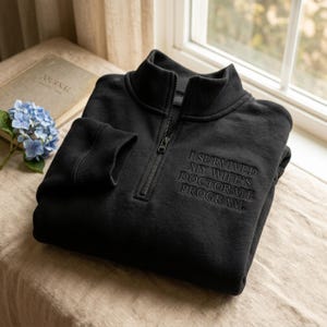May include: Black quarter-zip sweatshirt with the embroidered text "I Survived My Wife's Doctorate Program." The sweatshirt has a ribbed collar and cuffs. A journal and blue hydrangea flowers are in the background.