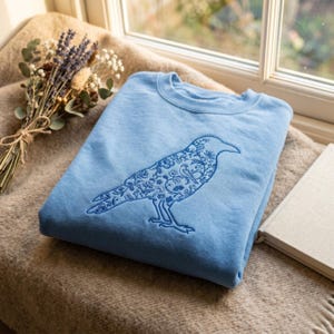 May include: A light blue sweatshirt featuring a detailed embroidered raven design. The raven is filled with floral and leaf patterns in a darker shade of blue. The sweatshirt is folded and resting on a neutral-toned blanket near a window.