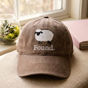 Puede incluir: Una gorra de béisbol marrón con un bordado de una oveja blanca con cara y patas negras. Debajo de la oveja, la palabra "Found." está bordada en hilo blanco. La gorra tiene una visera curva y una corona estructurada con detalles de costura visibles.