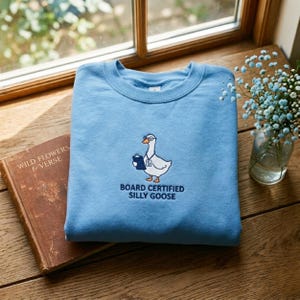 May include: A light blue sweatshirt with a crew neck. The front features an embroidered cartoon goose wearing glasses and holding a clipboard, with the text "BOARD CERTIFIED SILLY GOOSE" below. The sweatshirt is folded on a wooden surface.