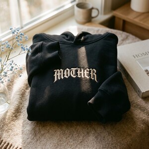 May include: A black hooded sweatshirt featuring the word MOTHER embroidered in a cream-colored gothic font across the center chest. The garment has long sleeves with ribbed cuffs and a hood, showcasing a clean, solid black fabric construction.