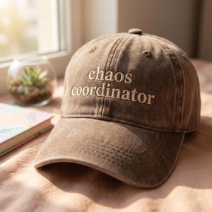 Peut inclure: Casquette de baseball marron avec le texte brodé "chaos coordinator" en blanc. La casquette est faite d'un tissu doux et usé et a une visière incurvée. La casquette est posée sur une surface rose.
