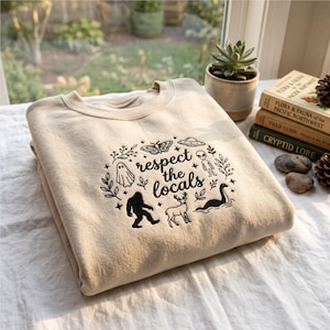 Puede incluir: Una sudadera beige de cuello redondo con un diseño bordado en negro que incluye el texto "respect the locals" rodeado de ilustraciones de un fantasma, una polilla, un OVNI, un alienígena, Bigfoot, un ciervo y el monstruo del lago Ness.