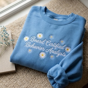 Könnte beinhalten: Hellblaues Sweatshirt mit der gestickten Aufschrift "Board Certified Behavior Analysts" und Gänseblümchen-Akzenten. Das Sweatshirt ist auf einer beigefarbenen, gewebten Oberfläche gefaltet, mit einem Buch und Trockenblumen im Hintergrund.
