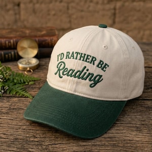 May include: A baseball cap with a cream-colored crown and a dark green brim. The cap features the embroidered phrase "I'd Rather Be Reading" in green script. The cap has a dark green button on top.
