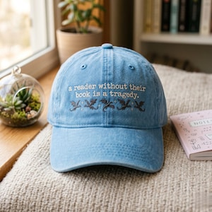 Op de afbeelding: Lichtblauwe baseballcap met geborduurde tekst: "a reader without their book is a tragedy." Onder de tekst staan geborduurde draken. De pet ligt op een beige oppervlak.