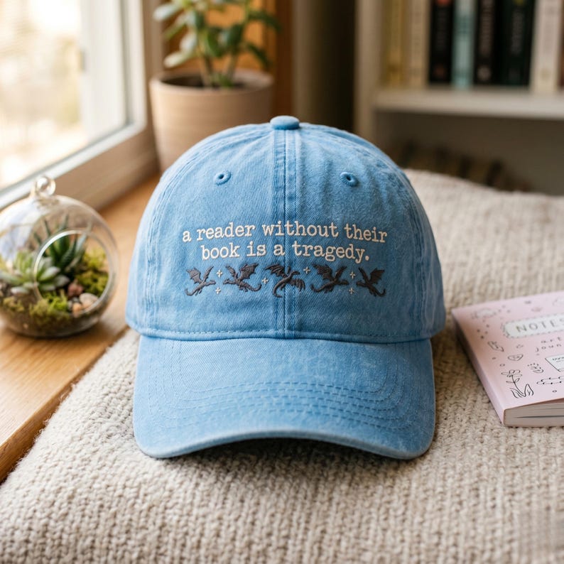 Op de afbeelding: Lichtblauwe baseballcap met geborduurde tekst: "a reader without their book is a tragedy." Onder de tekst staan geborduurde draken. De pet ligt op een beige oppervlak.