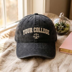 Peut inclure: Casquette de baseball noire avec l'inscription blanche "YOUR COLLEGE" en arc de cercle sur le devant. En dessous, un symbole de balance blanc. La casquette est posée sur une surface beige.